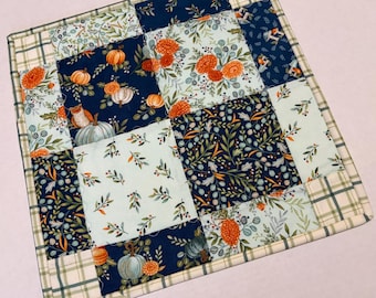 May include: A square quilted fabric mat featuring a patchwork design of alternating navy blue and light mint green squares. The fabric patterns include orange pumpkins, floral motifs, and leafy botanical prints, all bordered by a plaid fabric trim.