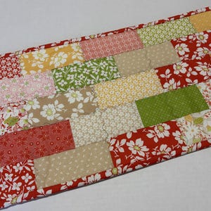May include: A rectangular fabric table runner featuring a patchwork design of various floral and geometric patterns in shades of red, green, yellow, pink, and beige. The runner is finished with a solid red fabric border along the edges.