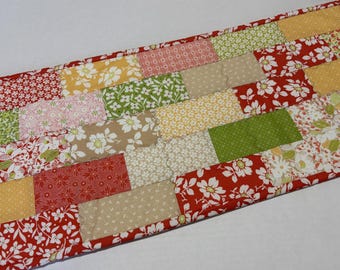 May include: A rectangular fabric table runner featuring a patchwork design of various floral and geometric patterns in shades of red, green, yellow, pink, and beige. The runner is finished with a solid red fabric border along the edges.
