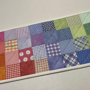 May include: A colorful patchwork quilt runner with a white border. The runner features a rainbow of square fabric patches in various patterns, including polka dots, gingham, and plaid. The quilt is machine-stitched.