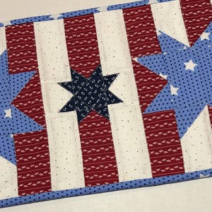 May include: A patriotic table runner featuring a repeating pattern of red and white stripes, interspersed with blue stars. The stars are filled with white dots and are set against a blue background. The runner is bordered with a blue fabric.