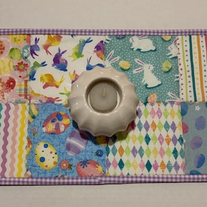 Może przedstawiać: Dekoracyjny świecznik w tematyce wielkanocnej z białą ceramiczną podstawą, umieszczony na patchworkowej podkładce. Podkładka posiada kolorowe kwadraty materiału z wzorami jajek wielkanocnych i kr&oacute;liczk&oacute;w, obramowane fioletową obw&oacute;dką w kratkę.