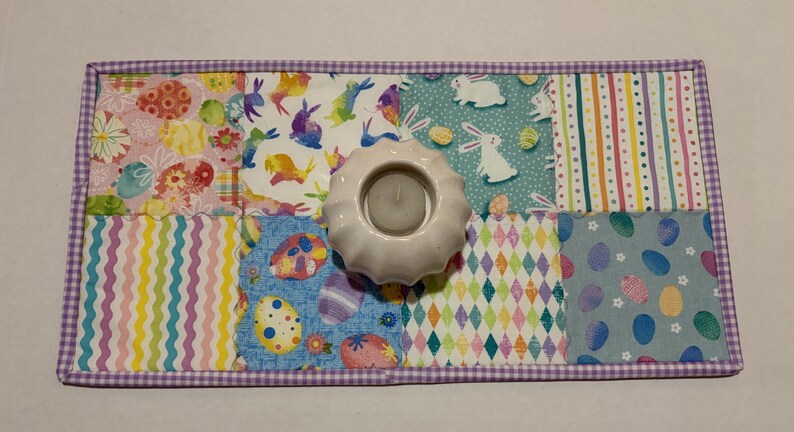 Może przedstawiać: Dekoracyjny świecznik w tematyce wielkanocnej z białą ceramiczną podstawą, umieszczony na patchworkowej podkładce. Podkładka posiada kolorowe kwadraty materiału z wzorami jajek wielkanocnych i kr&oacute;liczk&oacute;w, obramowane fioletową obw&oacute;dką w kratkę.