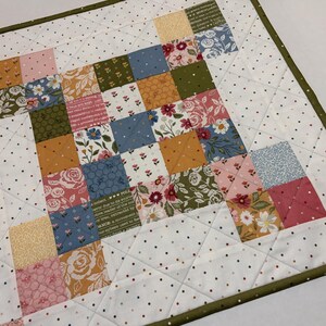 May include: A square patchwork quilt with a white background dotted with small, colorful polka dots. The quilt features a central design of colorful squares in shades of pink, blue, yellow, and green, with floral patterns. The quilt is trimmed with a green border.