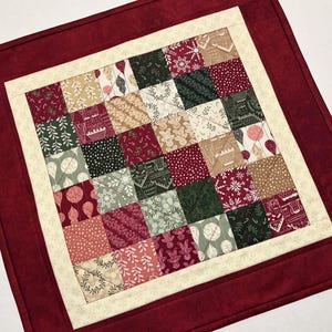 May include: A square quilted fabric mat featuring a patchwork center of small squares with various holiday-themed patterns in red, green, and cream. The center is framed by a cream-colored border and an outer border of solid dark red fabric.