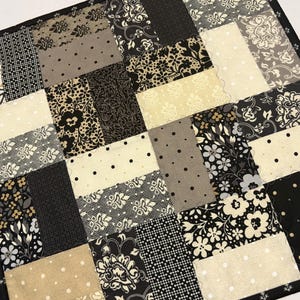 May include: A square patchwork quilt featuring a variety of rectangular fabric blocks in black, white, cream, and gray. The patterns include floral motifs, polka dots, and geometric grids, all bordered by a thin black fabric trim with white accents.
