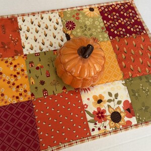 Peut inclure: Un set de table patchwork rectangulaire compos&eacute; de carr&eacute;s de tissu aux motifs automnaux dans des tons orange, jaune, vert et bordeaux, surmont&eacute; d'un r&eacute;cipient en c&eacute;ramique orange brillant en forme de citrouille avec un couvercle.