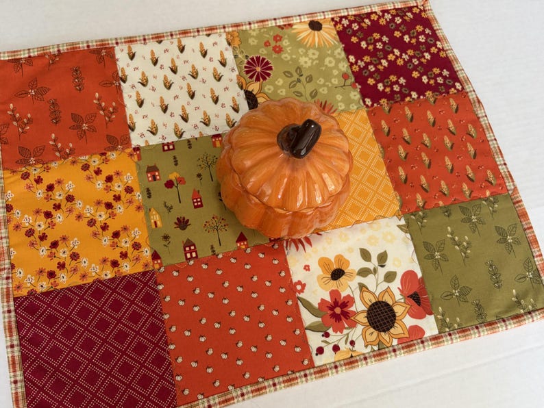 Peut inclure: Un set de table patchwork rectangulaire compos&eacute; de carr&eacute;s de tissu aux motifs automnaux dans des tons orange, jaune, vert et bordeaux, surmont&eacute; d'un r&eacute;cipient en c&eacute;ramique orange brillant en forme de citrouille avec un couvercle.