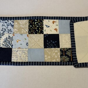 May include: A rectangular quilted table runner featuring a patchwork design of navy blue, light blue, cream, and white fabric squares. The patterns include birds, trees, and script text. The runner is finished with a navy blue and white striped binding.