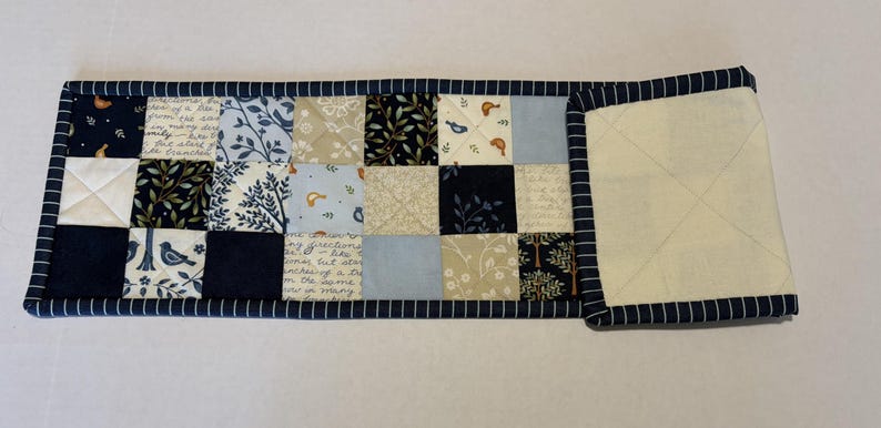 May include: A rectangular quilted table runner featuring a patchwork design of navy blue, light blue, cream, and white fabric squares. The patterns include birds, trees, and script text. The runner is finished with a navy blue and white striped binding.