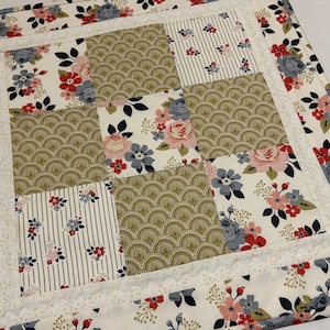 May include: A patchwork quilt featuring floral and geometric patterns in shades of white, pink, red, gray, and gold. The quilt has a white border with a delicate lace trim. The design includes roses, stripes, and a fan-like pattern.
