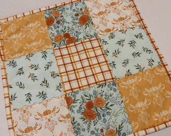 May include: A square fabric quilt featuring a patchwork design of nine alternating blocks. The patterns include orange pumpkin motifs on cream, light blue floral arrangements with orange flowers, and a central orange and white plaid square.