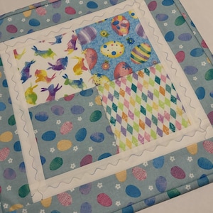 K&ouml;nnte beinhalten: Ein quadratischer, gesteppter Tischset zum Thema Ostern. Die Mitte zeigt vier Stoffquadrate mit Osterei- und Hasenmustern in Pastellfarben. Der Rand ist hellblau mit Eierdesigns, und die Kanten sind mit einem wei&szlig;en, wellenf&ouml;rmigen Stich versehen.