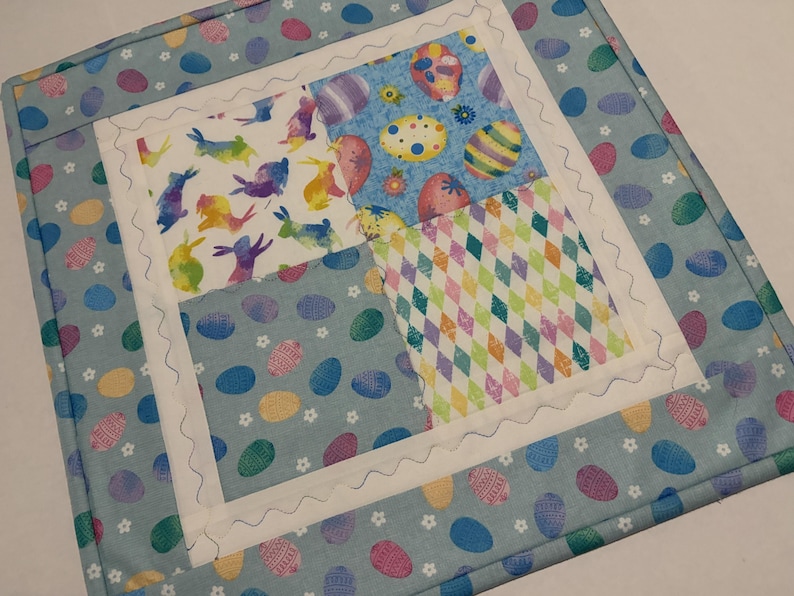 K&ouml;nnte beinhalten: Ein quadratischer, gesteppter Tischset zum Thema Ostern. Die Mitte zeigt vier Stoffquadrate mit Osterei- und Hasenmustern in Pastellfarben. Der Rand ist hellblau mit Eierdesigns, und die Kanten sind mit einem wei&szlig;en, wellenf&ouml;rmigen Stich versehen.