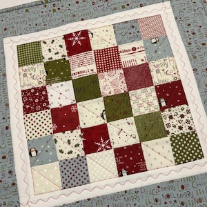May include: A square patchwork quilt featuring a grid of small fabric squares in red, green, white, and light blue. The patterns include snowflakes, penguins, and holiday text. A white border with wavy stitching frames the center, set on a light blue background with repeating Christmas words.
