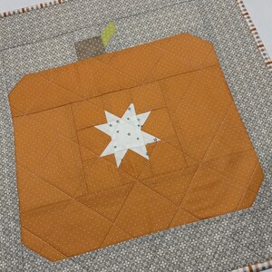 May include: A square quilted wall hanging featuring a central orange pumpkin block with a white star at its center. The pumpkin has a brown stem and a small green leaf. The background is a gray fabric with a white floral pattern and a plaid border.