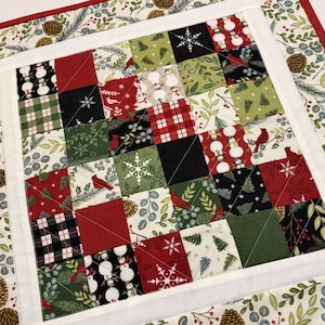 May include: A festive Christmas-themed quilted table runner. The center features a patchwork design with red, green, black, and white squares adorned with snowmen, snowflakes, and cardinal birds. The border has a white band and a red edge with a botanical print.