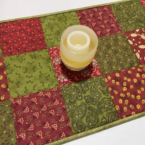 Puede incluir: Un camino de mesa acolchado con un dise&ntilde;o de patchwork en tonos rojos, verdes y crema. El camino de mesa presenta patrones florales y de cachemira. Un portavelas de vidrio con una vela blanca encima.