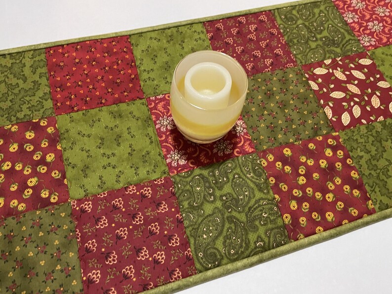 Puede incluir: Un camino de mesa acolchado con un dise&ntilde;o de patchwork en tonos rojos, verdes y crema. El camino de mesa presenta patrones florales y de cachemira. Un portavelas de vidrio con una vela blanca encima.
