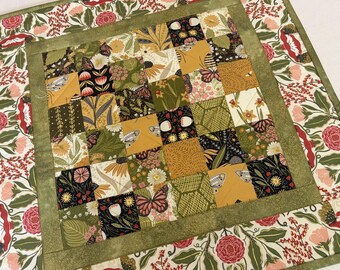 May include: A square fabric quilt featuring a central grid of patchwork squares in shades of mustard yellow, olive green, and black, decorated with butterfly and floral motifs. The center is framed by a solid olive green border and a floral outer border.