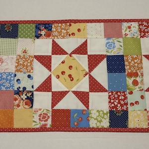 May include: A colorful quilted table runner with a patchwork design. The runner features squares of various patterns and colors, including red, white, blue, yellow, and green. Red star shapes with white centers and cherry patterns are also incorporated into the design.