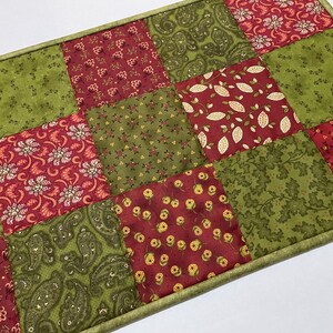Puede incluir: Un camino de mesa rectangular de patchwork con cuadrados de telas estampadas en rojo, verde y oliva. Las telas muestran dise&ntilde;os florales, de cachemira y de hojas. El camino de mesa est&aacute; bordeado con un borde verde.