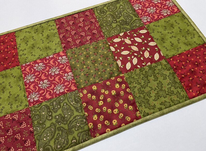 Puede incluir: Un camino de mesa rectangular de patchwork con cuadrados de telas estampadas en rojo, verde y oliva. Las telas muestran dise&ntilde;os florales, de cachemira y de hojas. El camino de mesa est&aacute; bordeado con un borde verde.