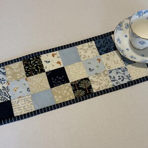 May include: A rectangular quilted table runner featuring a patchwork design of squares in shades of navy blue, light blue, cream, and white. The fabric squares display patterns of birds, trees, and script text, finished with a navy blue striped border.
