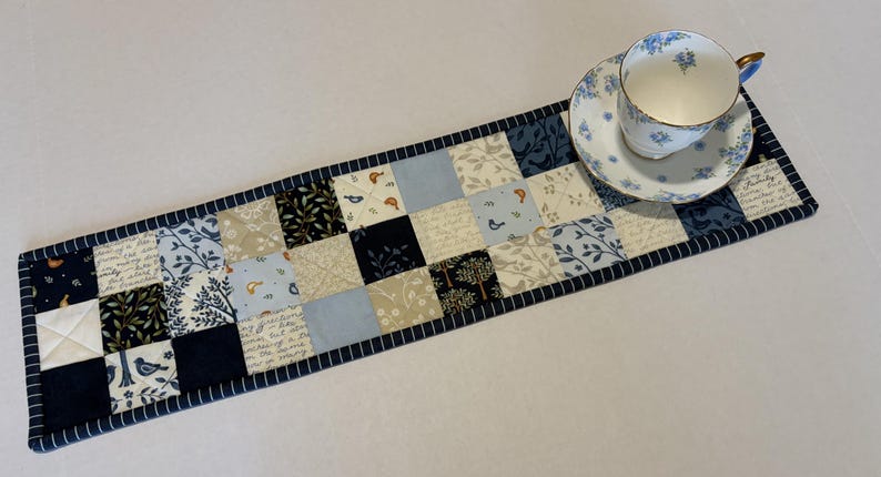 May include: A rectangular quilted table runner featuring a patchwork design of squares in shades of navy blue, light blue, cream, and white. The fabric squares display patterns of birds, trees, and script text, finished with a navy blue striped border.