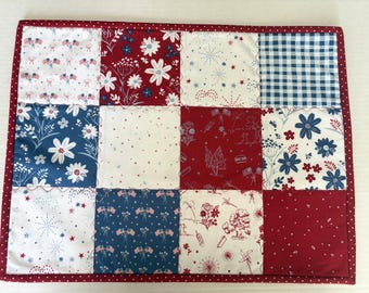 May include: A rectangular quilted placemat featuring a patchwork design of twelve squares in red, white, and blue. The fabric patterns include floral motifs, gingham checks, and small American flag illustrations. It is finished with a red polka dot border.