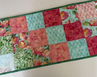 Peut inclure: Un chemin de table rectangulaire composé de panneaux de tissu carrés en patchwork. Le motif présente des imprimés floraux avec des roses roses, rouges et orange, un feuillage vert et des formes géométriques bleu clair, bordé d'un liseré vert.