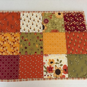 Peut inclure: Un set de table rectangulaire en tissu compos&eacute; d'un patchwork de douze carr&eacute;s. Le motif pr&eacute;sente des &eacute;l&eacute;ments sur le th&egrave;me de l'automne comme des tournesols, des &eacute;pis de ma&iuml;s, des petites maisons et des formes g&eacute;om&eacute;triques aux tons orange, jaune, rouge et vert olive.