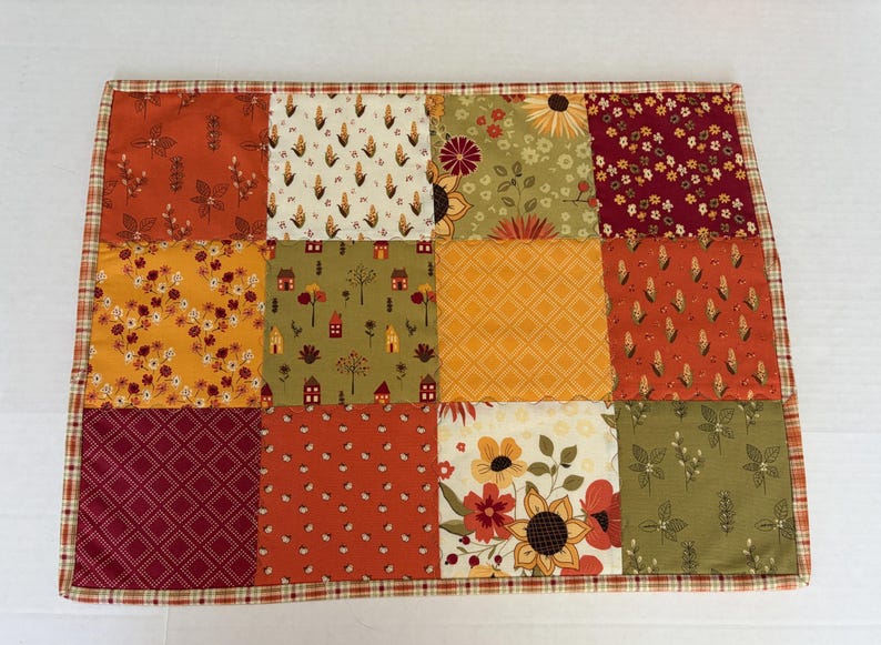 Peut inclure: Un set de table rectangulaire en tissu compos&eacute; d'un patchwork de douze carr&eacute;s. Le motif pr&eacute;sente des &eacute;l&eacute;ments sur le th&egrave;me de l'automne comme des tournesols, des &eacute;pis de ma&iuml;s, des petites maisons et des formes g&eacute;om&eacute;triques aux tons orange, jaune, rouge et vert olive.