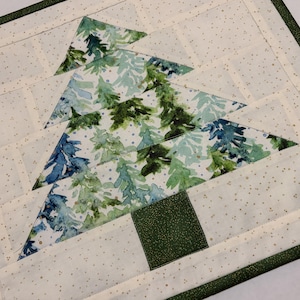 May include: A quilted Christmas tree design on a cream-colored background with small gold dots. The tree is made of green and blue watercolor-style fabric. The edges are finished with a dark green border.