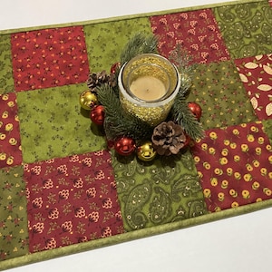 Puede incluir: Un camino de mesa navide&ntilde;o festivo con un dise&ntilde;o de patchwork en rojo y verde. Una vela en un portavelas dorado est&aacute; rodeada de agujas de pino, pi&ntilde;as y adornos rojos y dorados. El camino de mesa mide aproximadamente 30 cm de ancho.