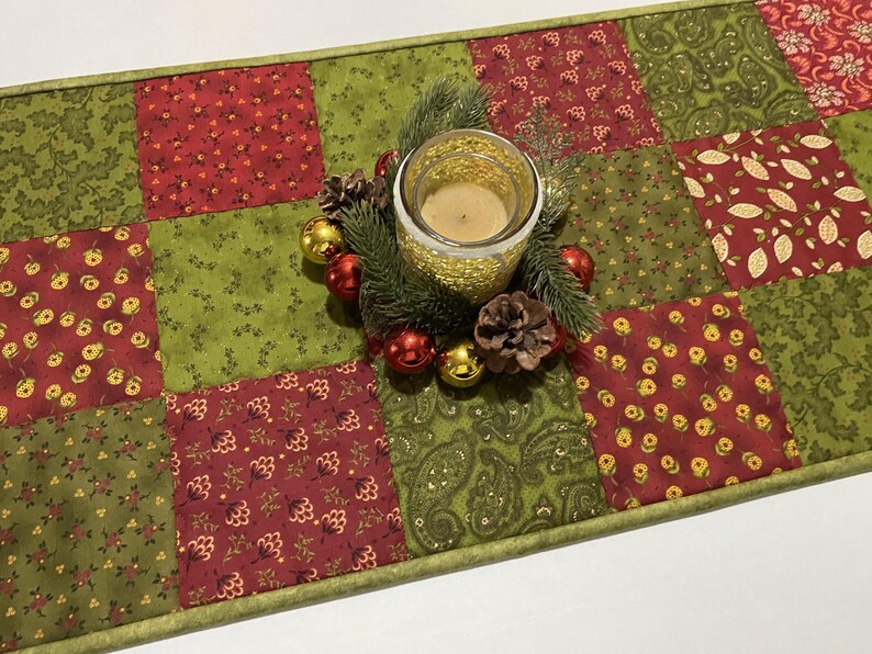 Puede incluir: Un camino de mesa navide&ntilde;o festivo con un dise&ntilde;o de patchwork en rojo y verde. Una vela en un portavelas dorado est&aacute; rodeada de agujas de pino, pi&ntilde;as y adornos rojos y dorados. El camino de mesa mide aproximadamente 30 cm de ancho.