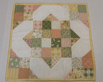 May include: A square patchwork quilt featuring a central star pattern composed of small fabric squares in shades of sage green, soft peach, yellow, and white. The design includes floral, gingham, and striped prints, finished with a yellow border.