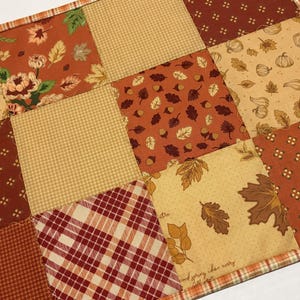 May include: A rectangular patchwork placemat featuring a variety of fabric squares in shades of orange, brown, and cream. The patterns include autumn leaves, pumpkins, acorns, small geometric shapes, and a red and white plaid design with a border.
