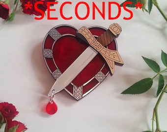 May include: A red heart-shaped brooch with a silver dagger piercing through it. The dagger has a gold handle and a brown wrapped hilt. A red teardrop hangs from the bottom of the dagger. The brooch is surrounded by a silver Celtic knot design. The text *SECONDS* is written in red letters above the brooch.