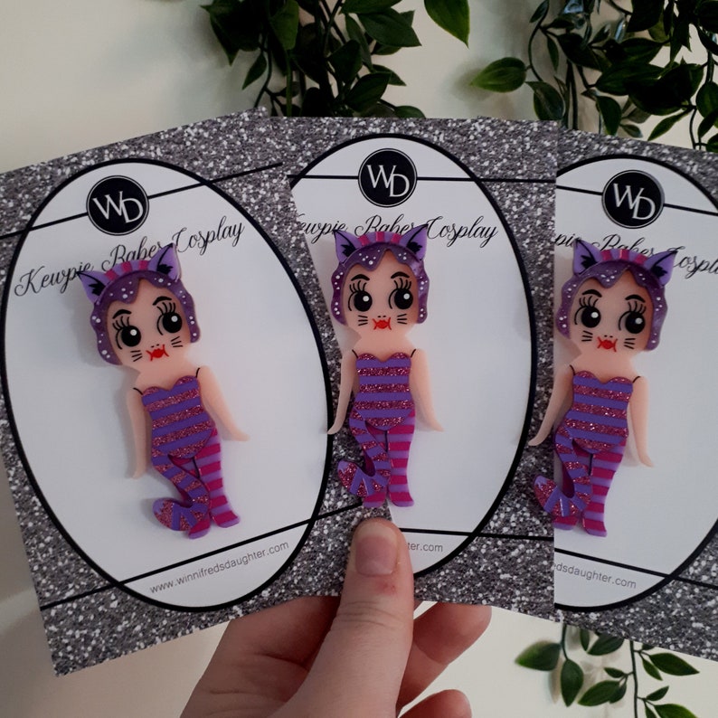 May include: Three purple and pink Kewpie doll pins with cat ears and striped outfits. The pins are on cards with the text "Kewpie Rabe Cosplay" and a website address.