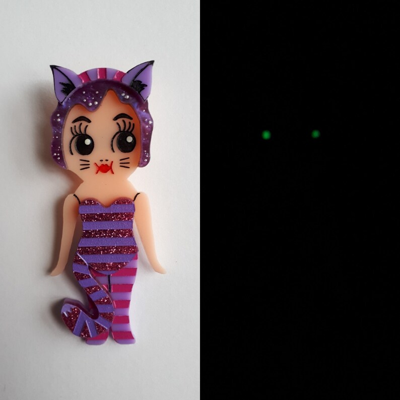 May include: A purple and pink cat-themed brooch with a striped dress and a cat face. The brooch glows in the dark with green eyes.