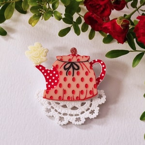 May include: A brooch shaped like a teapot with a peach-coloured body featuring red polka dots, a red and white polka-dot spout and handle, a black bow detail, a small red knob on the lid, and a pale yellow steam puff, resting on a white lace doily.