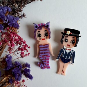 May include: Two colorful, hand-painted resin brooches featuring a purple cat with a pink and purple striped dress and a woman with a black and white striped dress and a black hat with a white flower.