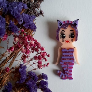 May include: Purple and pink cat-themed brooch with a glittery design. The brooch features a cartoon cat with a pink and purple striped dress and a pink bow.