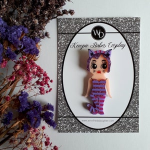 May include: Purple and pink Kewpie doll pin with a cat costume. The doll has a pink and white striped dress, purple cat ears, and a pink tail. The pin is on a white card with the text "Kewpie Babes Cosplay" and a website address.