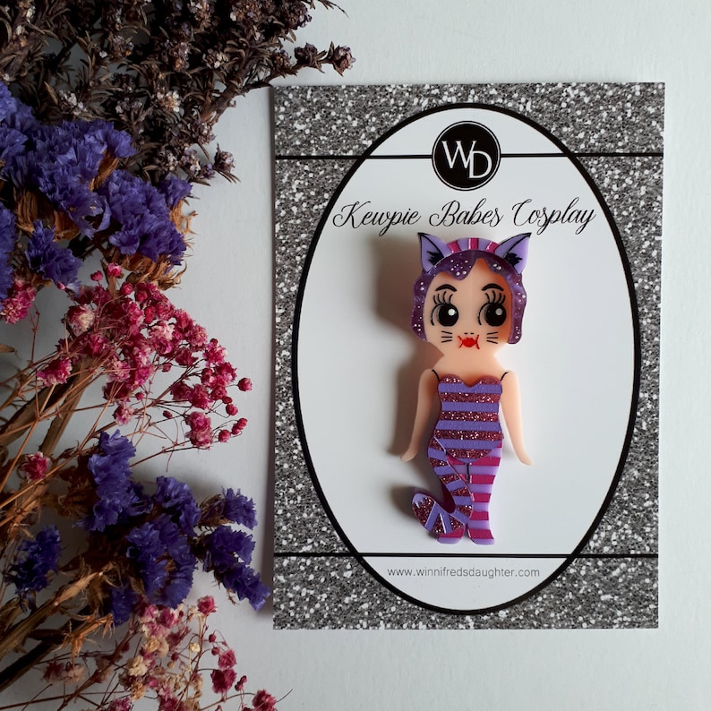 May include: Purple and pink Kewpie doll pin with a cat costume. The doll has a pink and white striped dress, purple cat ears, and a pink tail. The pin is on a white card with the text "Kewpie Babes Cosplay" and a website address.