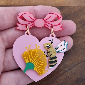 Op de afbeelding: Roze hartvormige oorbellen met een gele bloem en een bijenontwerp. De oorbellen hebben een roze strik aan de bovenkant. De bij heeft zwart-gouden strepen en een lichtblauwe vleugel.
