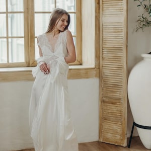 May include: A white satin nightgown with lace trim and a matching robe. The gown has a V-neck and delicate lace detailing on the bodice and sleeves. The model is wearing nude high heels.