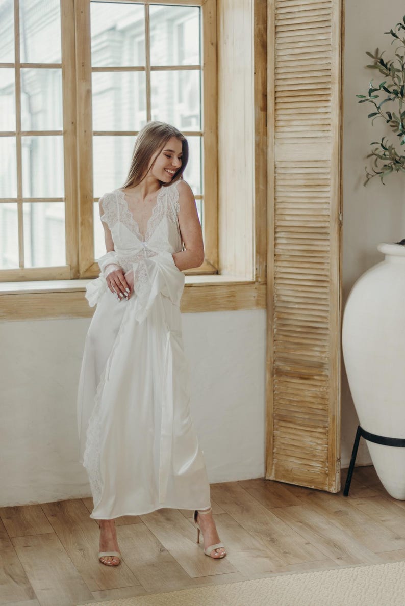May include: A white satin nightgown with lace trim and a matching robe. The gown has a V-neck and delicate lace detailing on the bodice and sleeves. The model is wearing nude high heels.