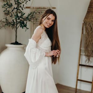 May include: A white satin bridal robe and gown with lace detailing. The gown has a V-neckline and a flowing skirt. The robe has wide sleeves. The image is taken indoors with a decorative plant and ladder in the background.